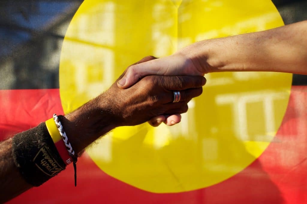 Black and white people shaking hands in front of the Aboriginal Flag